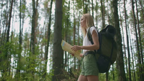 Young-caucasian-woman-looking-for-direction-on-a-map-while-hiking-in-the-forest.-Happy-girl-while-hiking-in-nature-and-orienteering-with-help-of-a-map